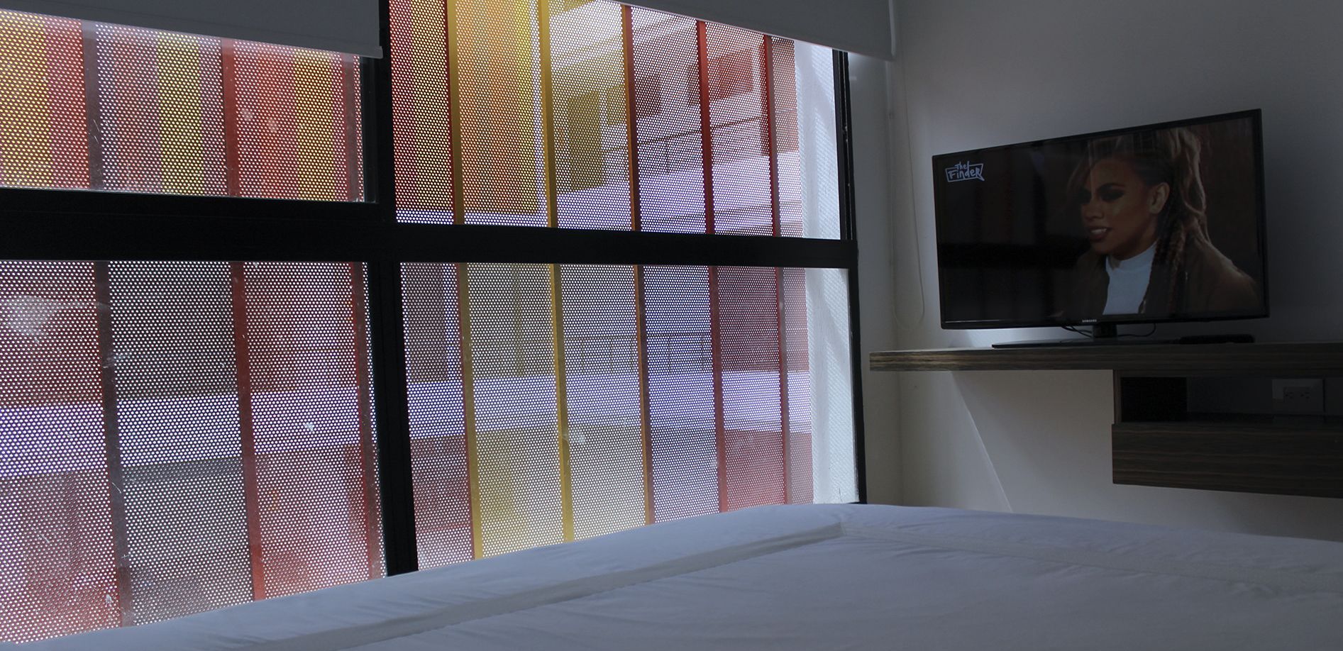 A hotel room with a bed, a TV mounted on the wall, and a window with a red and yellow patterned blind.