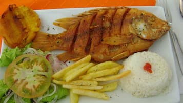 A plate with a grilled fish, French fries, a slice of tomato, lettuce, and white rice.