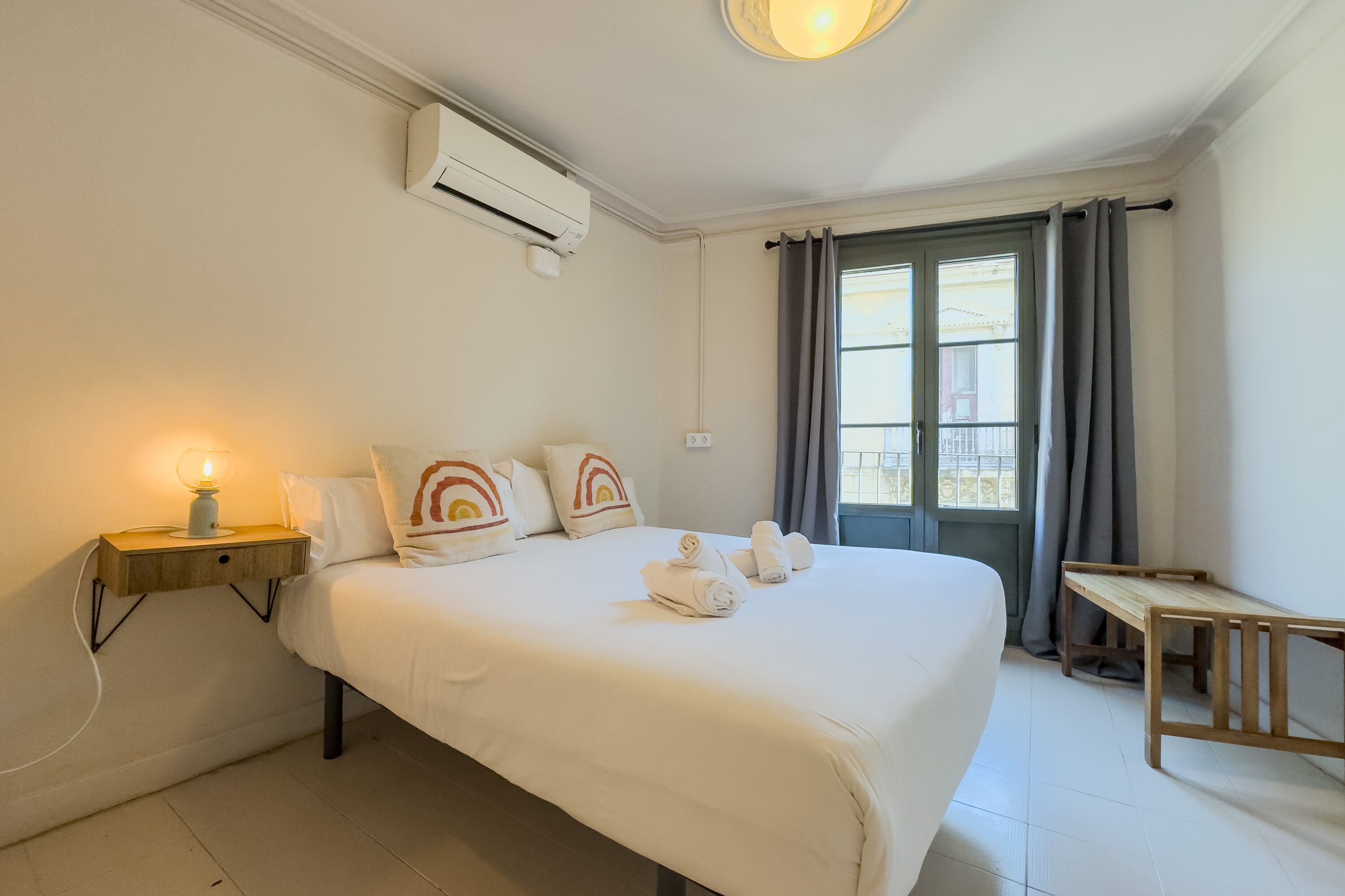 Een hotelkamer met een bed, nachtkastje, lamp, airconditioner en een raam met gordijnen.