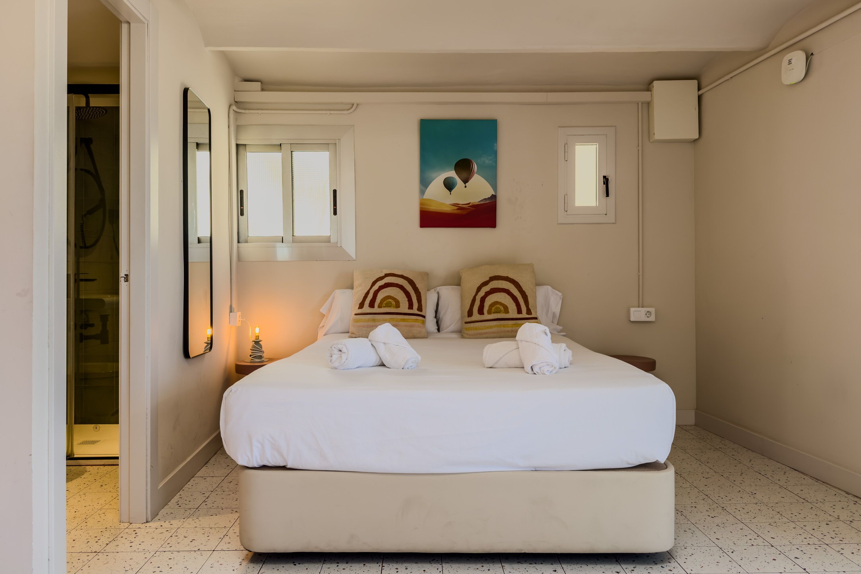A neatly made bed with towels, a painting of a person with hot air balloons, and a bathroom with a shower.
