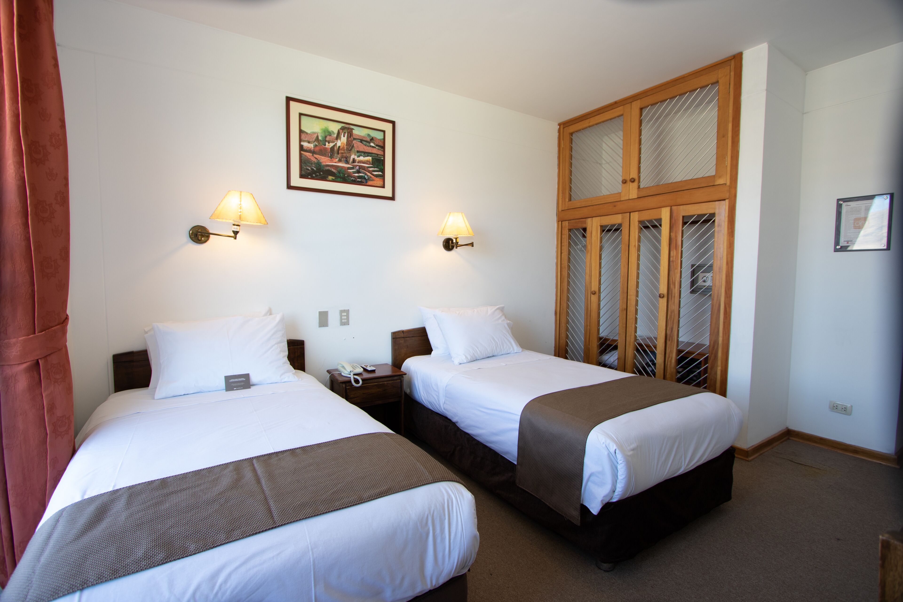 Une chambre d’hôtel avec deux lits, une armoire en bois, un tableau au mur et deux lampes fixées au mur.