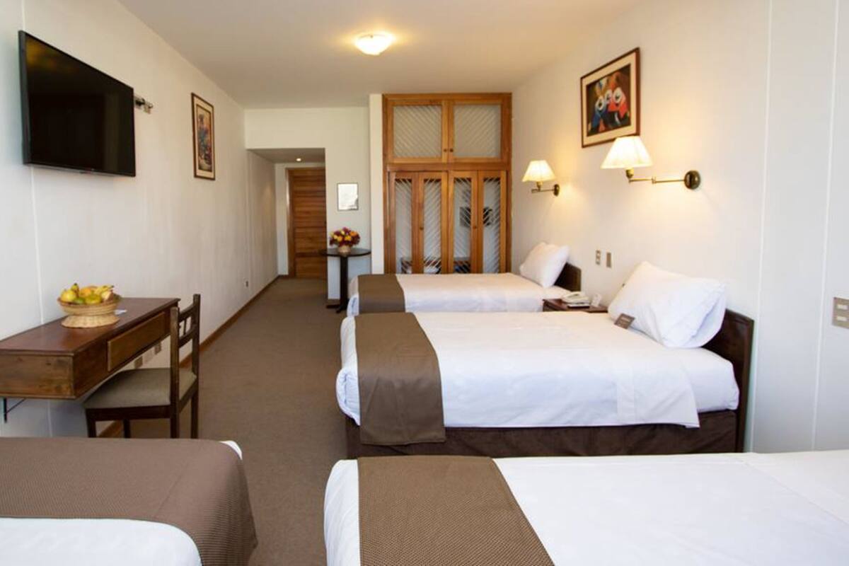 Quarto de hotel com duas camas, uma secretária com uma fruteira, uma televisão e uma porta de madeira com painéis de vidro.
