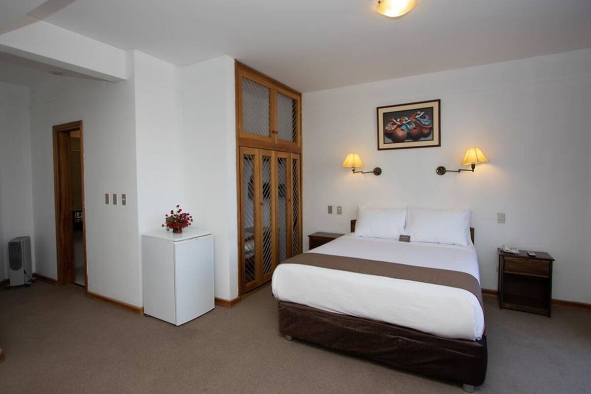 Quarto com uma cama grande, um frigorífico pequeno branco, um guarda-roupa de madeira e duas luzes de parede.