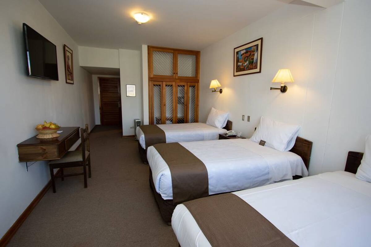 Quarto de hotel com três camas, uma escrivaninha com uma cesta de frutas, uma televisão e uma porta que dá para outro quarto.