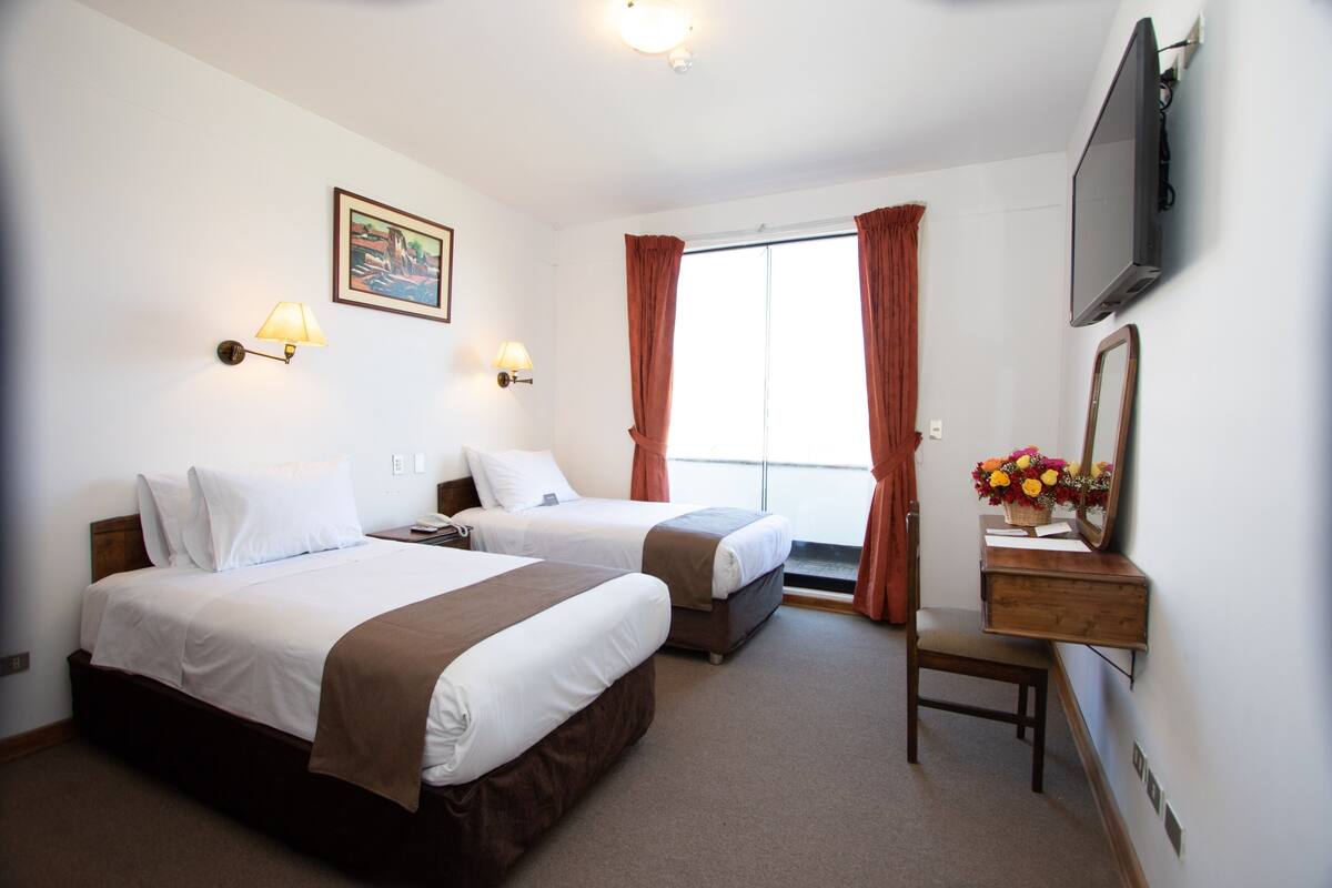 Quarto de hotel com duas camas, uma escrivaninha com espelho, televisão e janela com cortinas.
