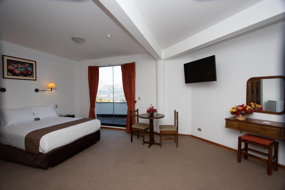 Quarto de hotel com cama, escrivaninha e cadeira, pequena mesa com um vaso de flores, televisão e janela com cortinas.