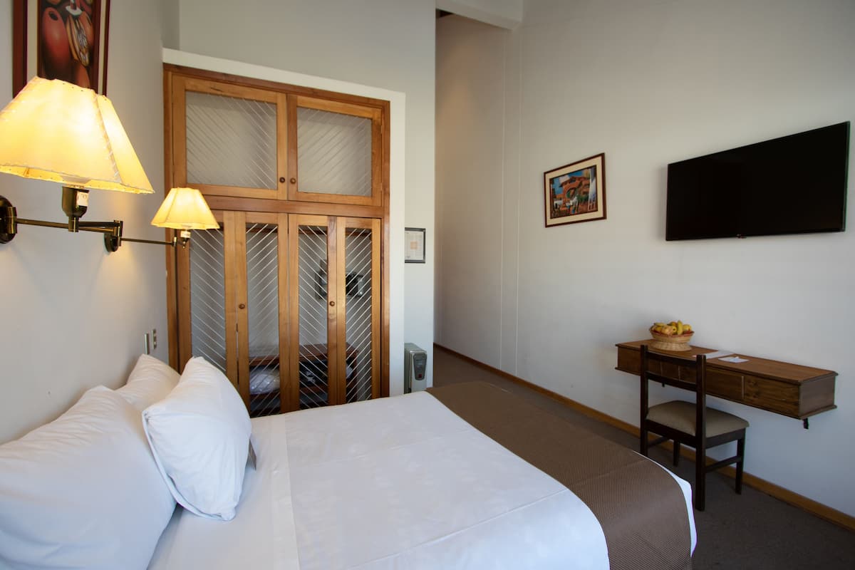 Quarto de hotel com cama, escrivaninha, cadeira, televisão e guarda-roupa com espelho.