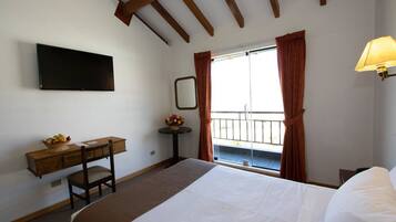 Habitación de hotel con cama, escritorio con silla, televisión en la pared, ventana con cortinas y lámpara.