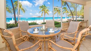A round glass table with chairs, a view of the beach, and palm trees. - The Colonial Club, Oceanfront Condos (Seven Mile Beach)