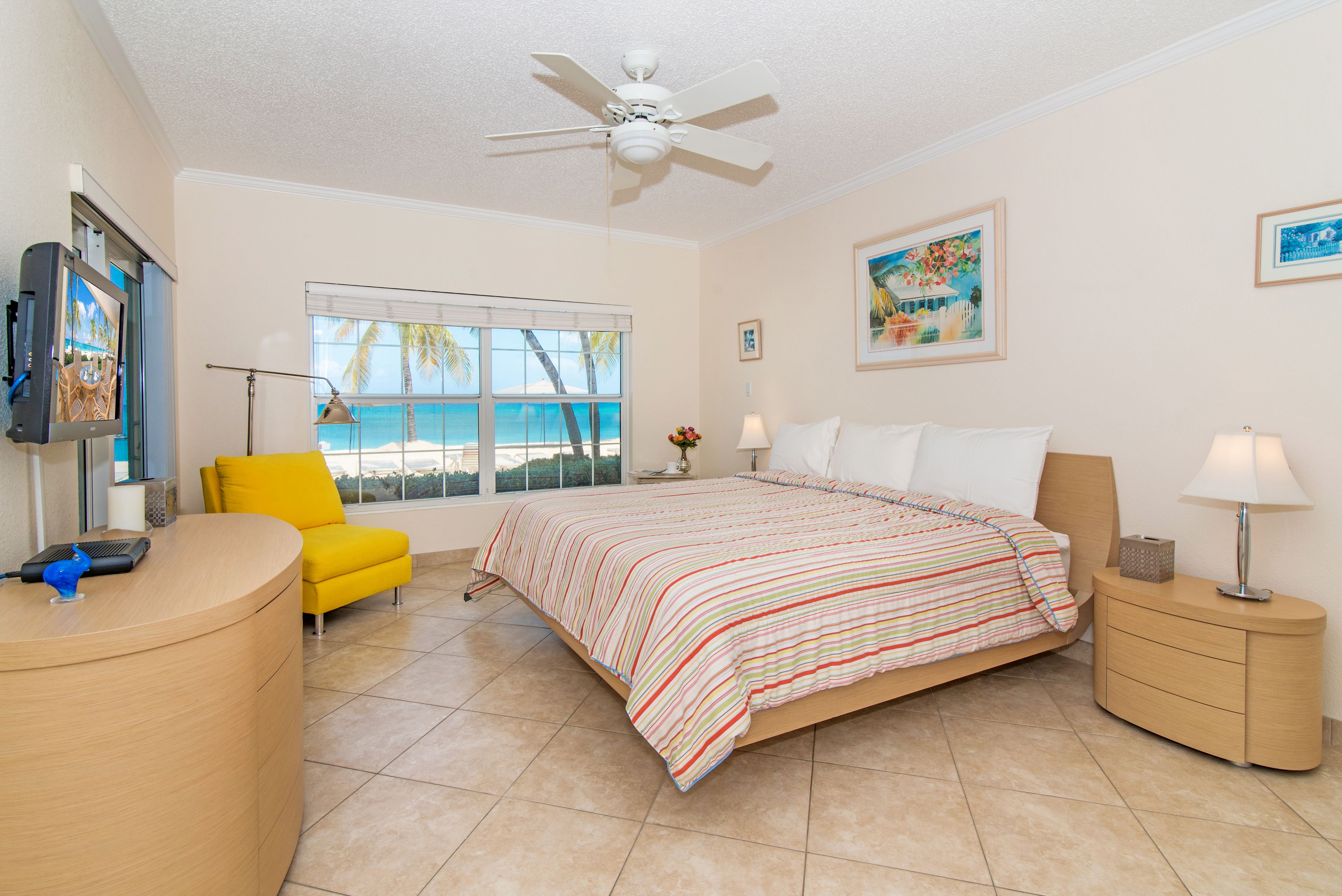 A hotel room with a bed, a desk, a chair, a window with a view of the beach, and a ceiling fan.