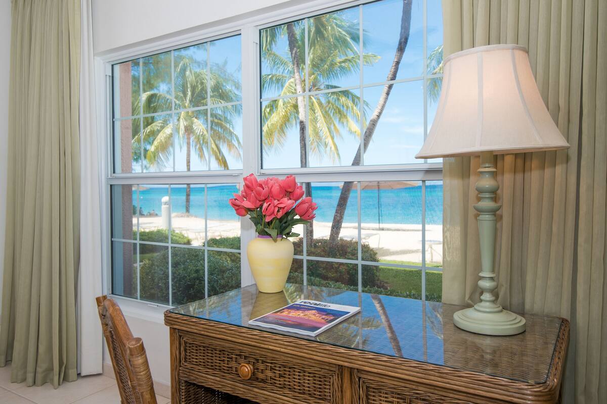 A room with a view of the beach, a wicker table with a lamp, a vase of flowers, and a magazine.