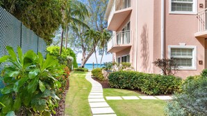 A well-maintained garden with a stone pathway leading to a building with balconies overlooking a body of water. - The Colonial Club, Oceanfront Condos (Seven Mile Beach)