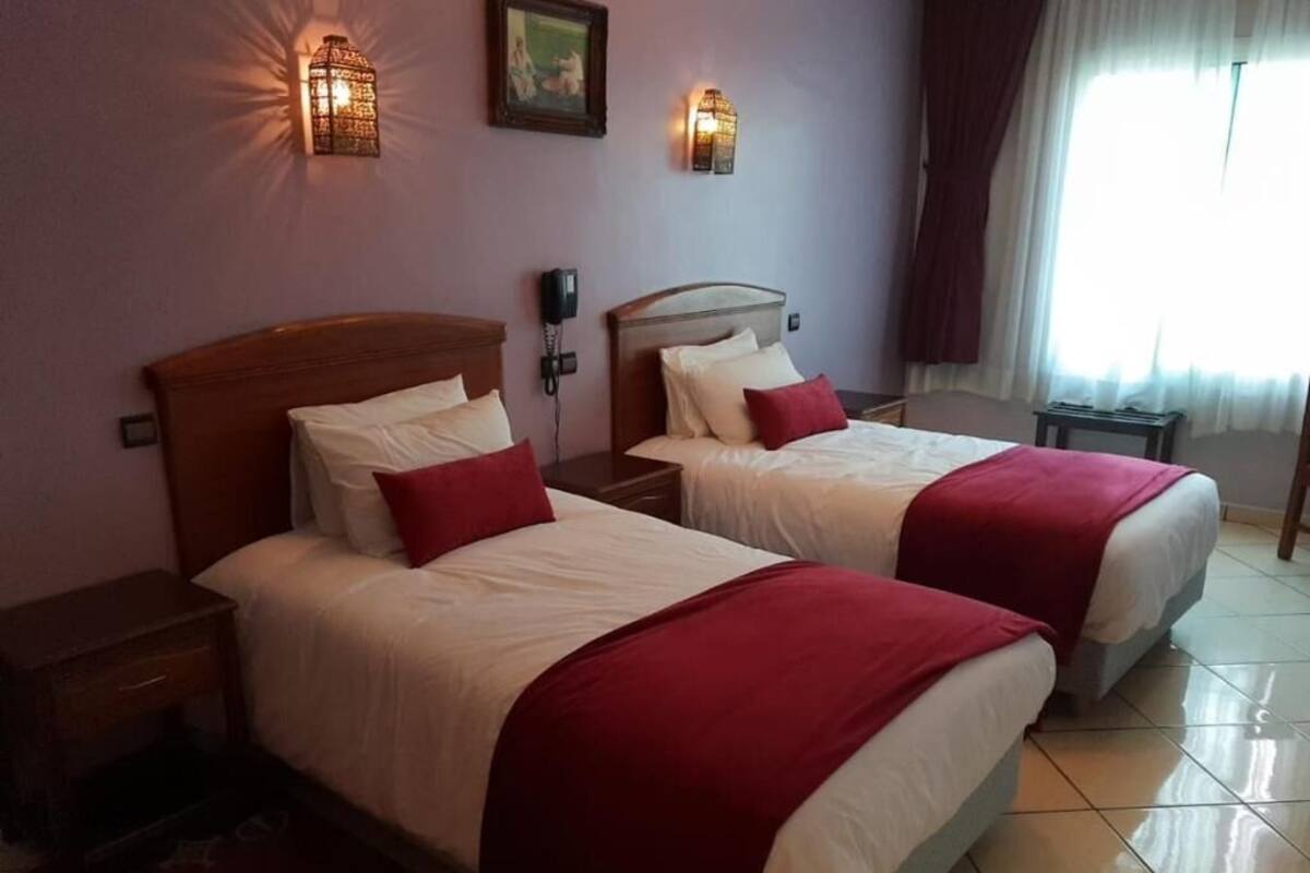 A hotel room with two beds, each with a wooden headboard, white bedding, and red pillows. There are wall-mounted light fixtures and a window with curtains.