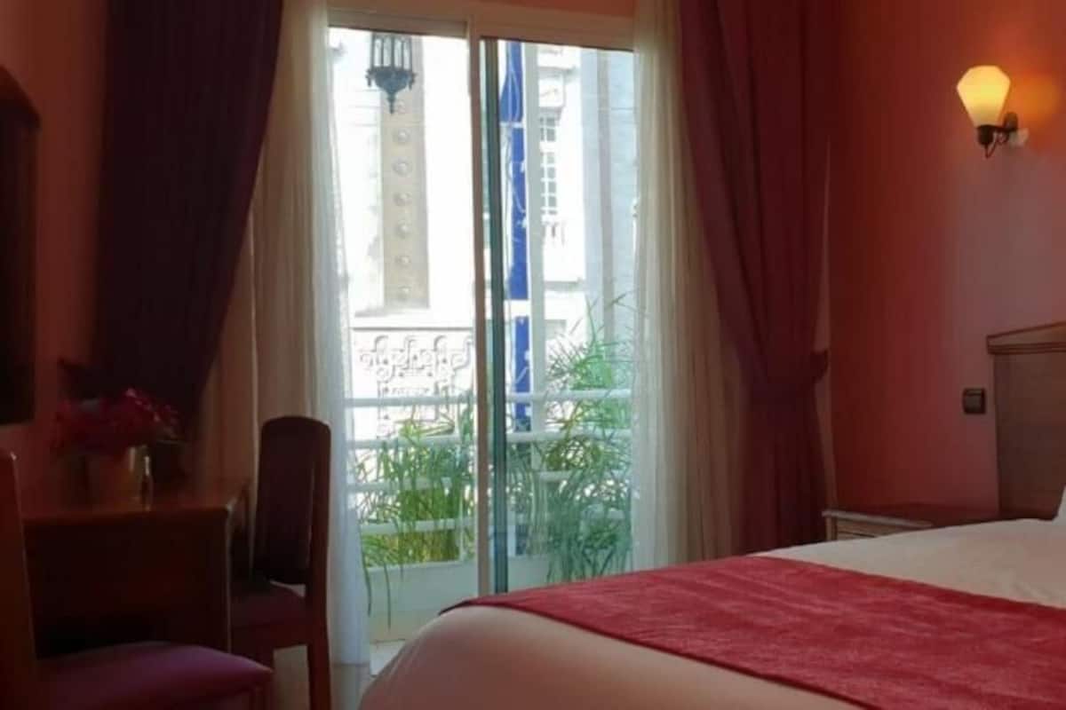 A hotel room with a bed, a desk, a chair, a window with curtains, and a wall-mounted light fixture.