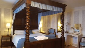 Una cama con dosel y cortinas, una mesita de noche con lámpara, una silla, un escritorio con espejo y un teléfono.