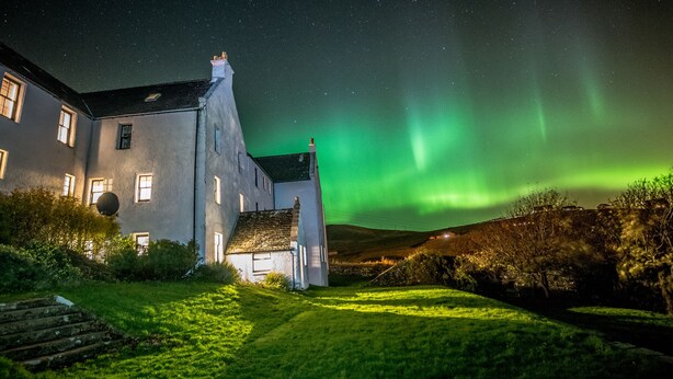 Un edificio con varias ventanas, un área verde en primer plano y un cielo nocturno con aurora boreal verde.