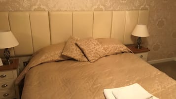 Una cama bien hecha con una colcha color beige, dos almohadas y una toalla blanca doblada. Hay dos mesitas de noche con lámparas y un aparador con una lámpara.
