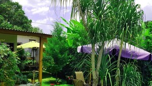 Un jardín exuberante con diversas plantas y árboles, un pequeño patio con mesa y sillas, y un edificio con tejado amarillo.