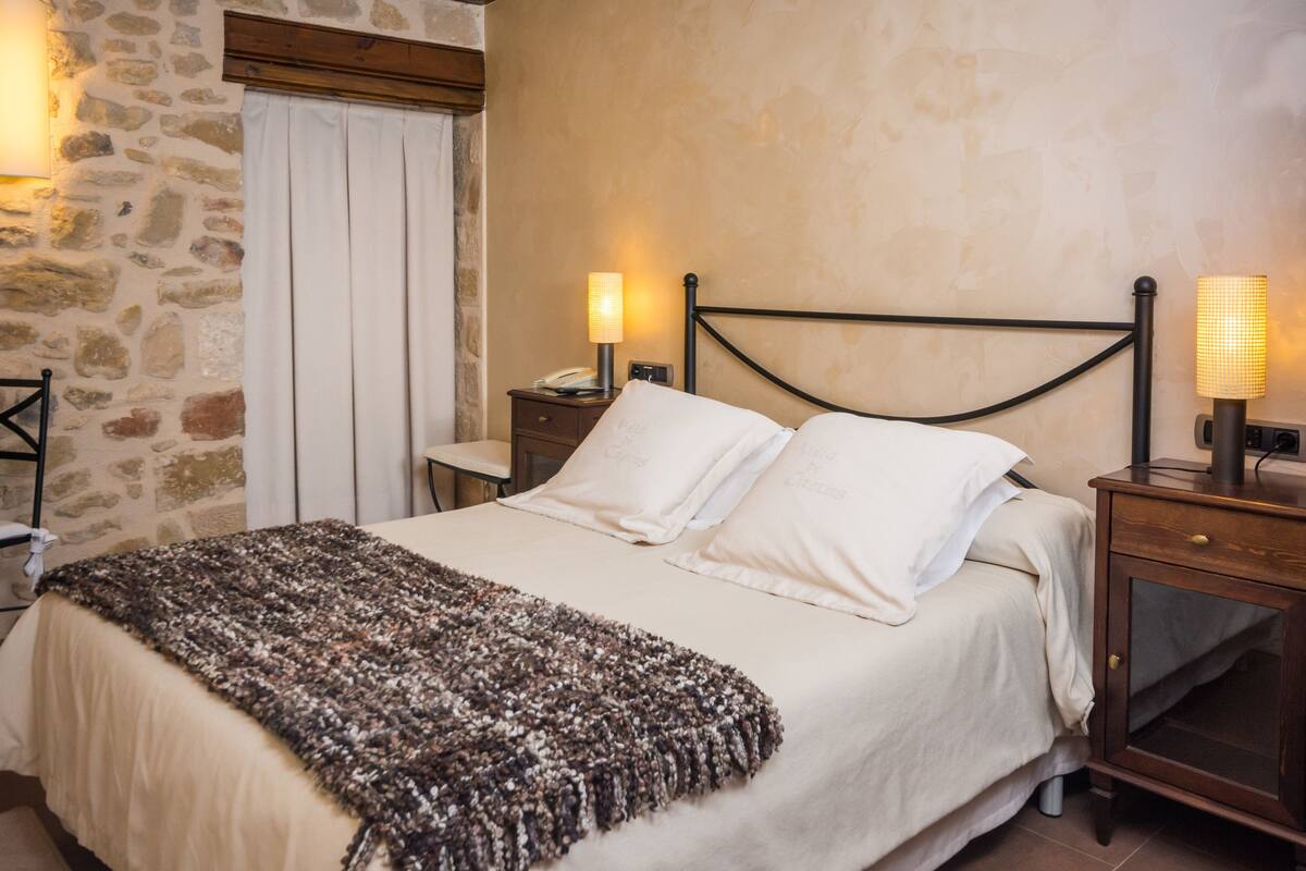 A neatly made bed with a patterned throw, a bedside table with a lamp, and a stone wall.