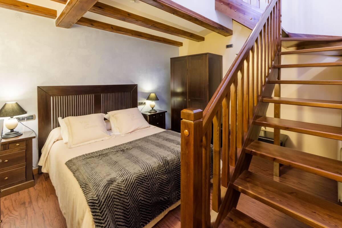 A bedroom with a wooden bed, a nightstand with a lamp, a wardrobe, and a wooden staircase.