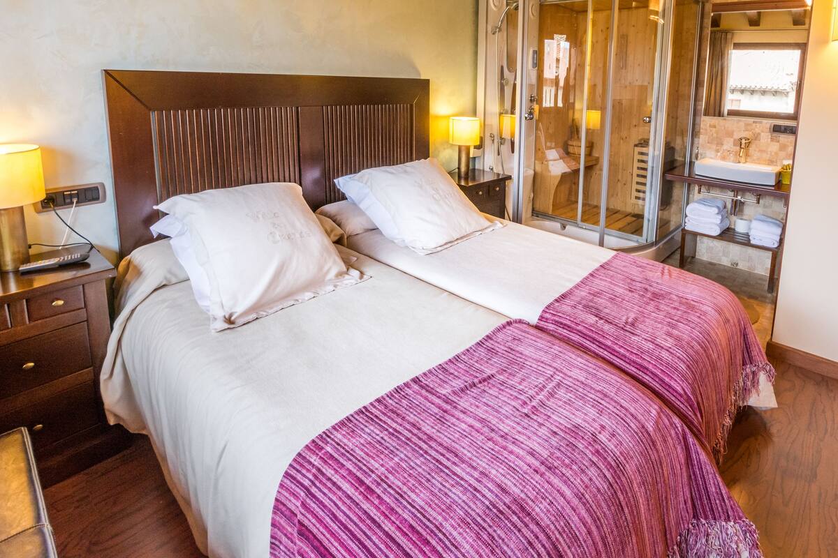 A double bed with a wooden headboard, two bedside tables with lamps, a glass-enclosed shower, and a wooden cabinet with towels.