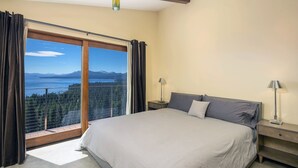 A bedroom with a large bed, a nightstand, a lamp, and a view of a lake and mountains through a sliding door. - Beautiful View by Hauserman Rentals (Tahoe City)
