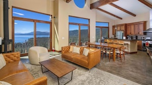 A spacious living area with a large sofa, a coffee table, and a dining table with chairs. There are large windows offering a view of mountains and a body of water. The kitchen area is visible with wooden cabinets and a stove. - Beautiful View by Hauserman Rentals (Tahoe City)