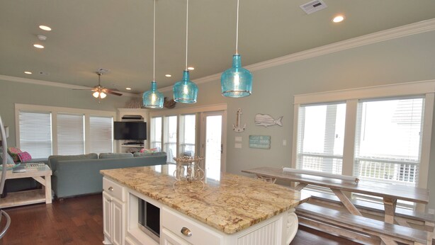 A kitchen with a granite countertop, white cabinetry, and a wooden island. There are hanging blue pendant lights, a ceiling fan, and a large window with blinds. - Luxury in Crystal Beach~ 4/3 ~ Beautiful Beach Views~Direct beach access (Crystal Beach)