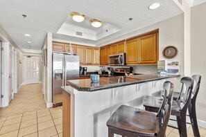 A kitchen with wooden cabinets, a stainless steel refrigerator, a microwave, and a sink. There is a clock on the wall and a bar stool in front of the counter. - Seawind 1308 by Liquid Life (Gulf Shores)