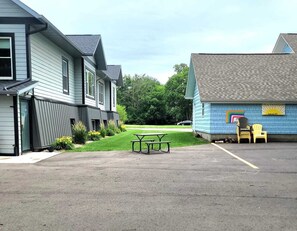 Un aparcamiento con mesa y sillas de picnic, un pequeño edificio con fachada azul y una casa con revestimiento gris.