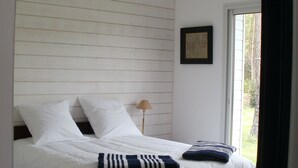 Ein Bett mit weißer Bettwäsche und einer Streifen-Decke, eine Nachttischlampe und ein Fenster mit Blick auf Bäume.