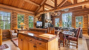 L’intérieur d’une cabane en rondins, avec une grande table à manger en bois, un îlot de cuisine et une cheminée en pierre.