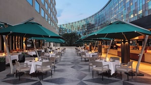 Un espace de restauration en plein air avec des tables et des chaises, des parasols verts et un bâtiment moderne en arrière-plan.