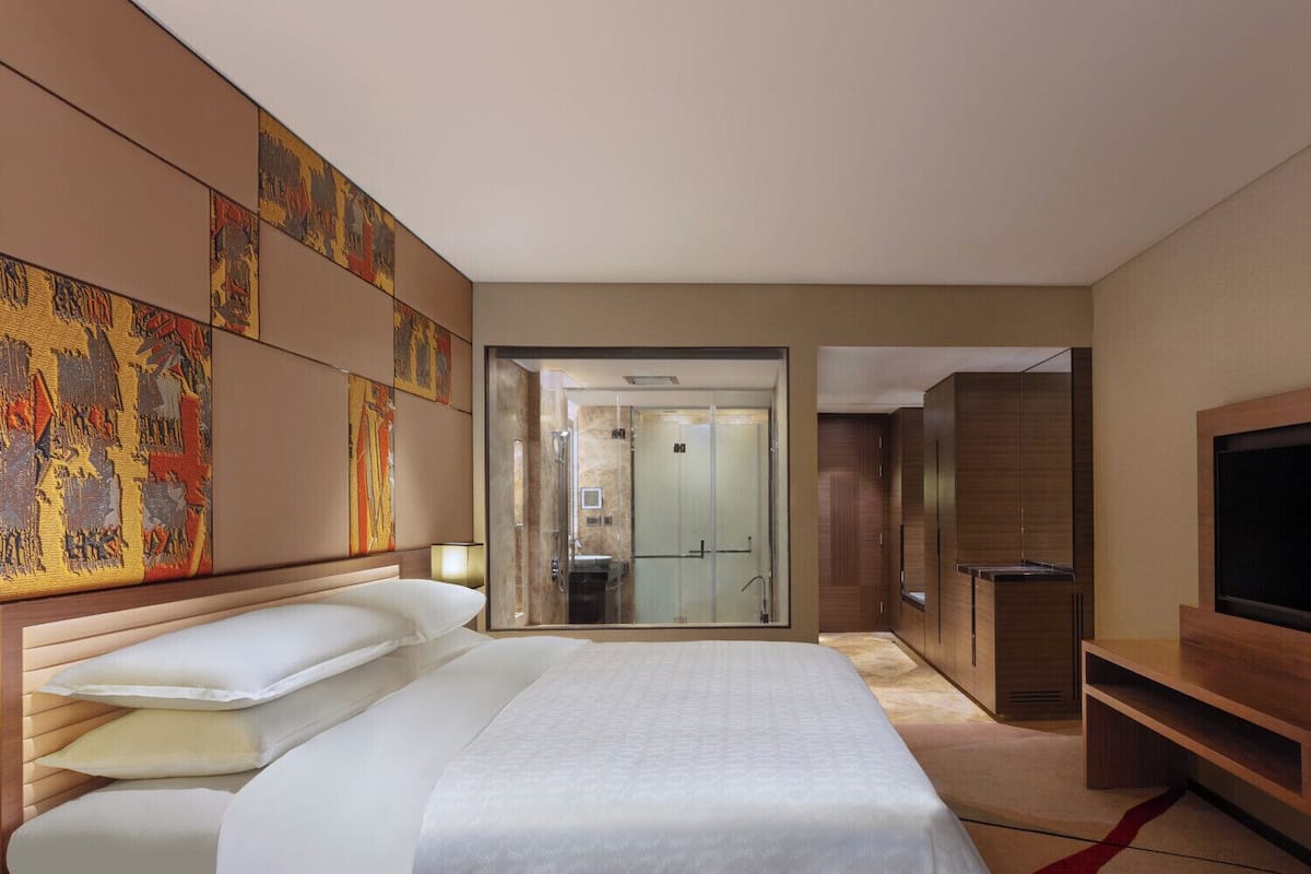 Habitación de hotel con una cama grande, televisión, armario y baño visible a través de una puerta de vidrio.
