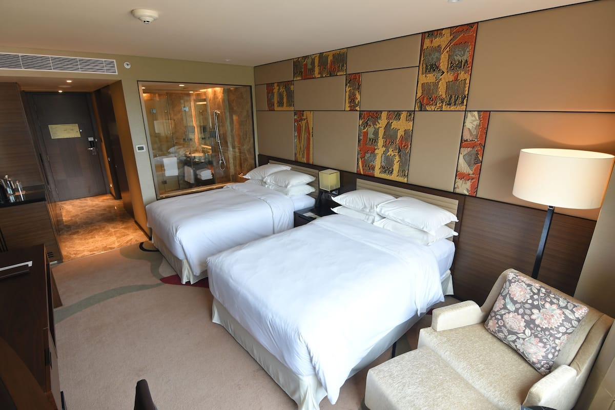 Habitación de hotel con dos camas, una zona de estar con una silla, un espejo grande y obras de arte en las paredes.