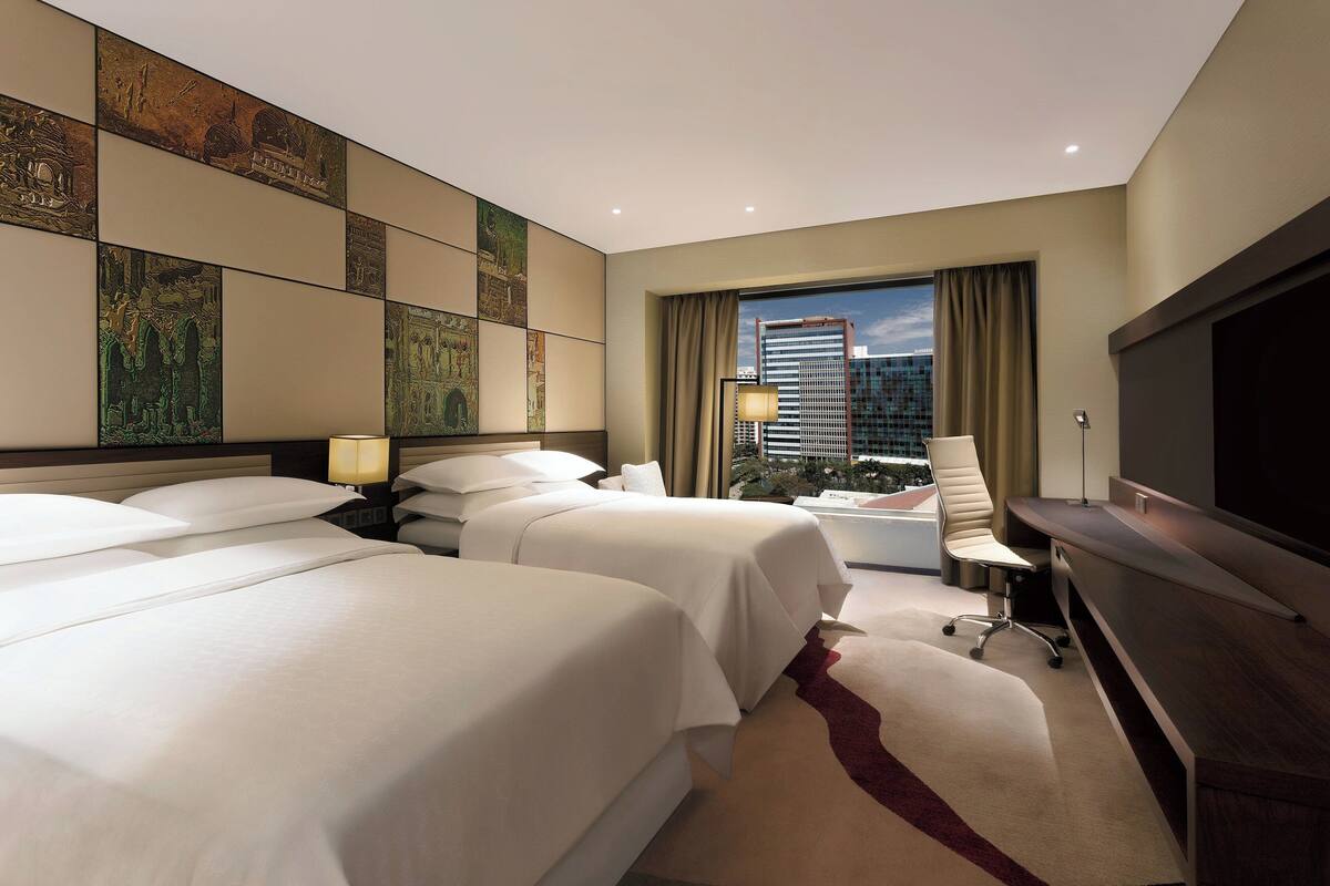 Habitación de hotel con dos camas, un escritorio, una silla, un televisor y una ventana con vista a la ciudad.