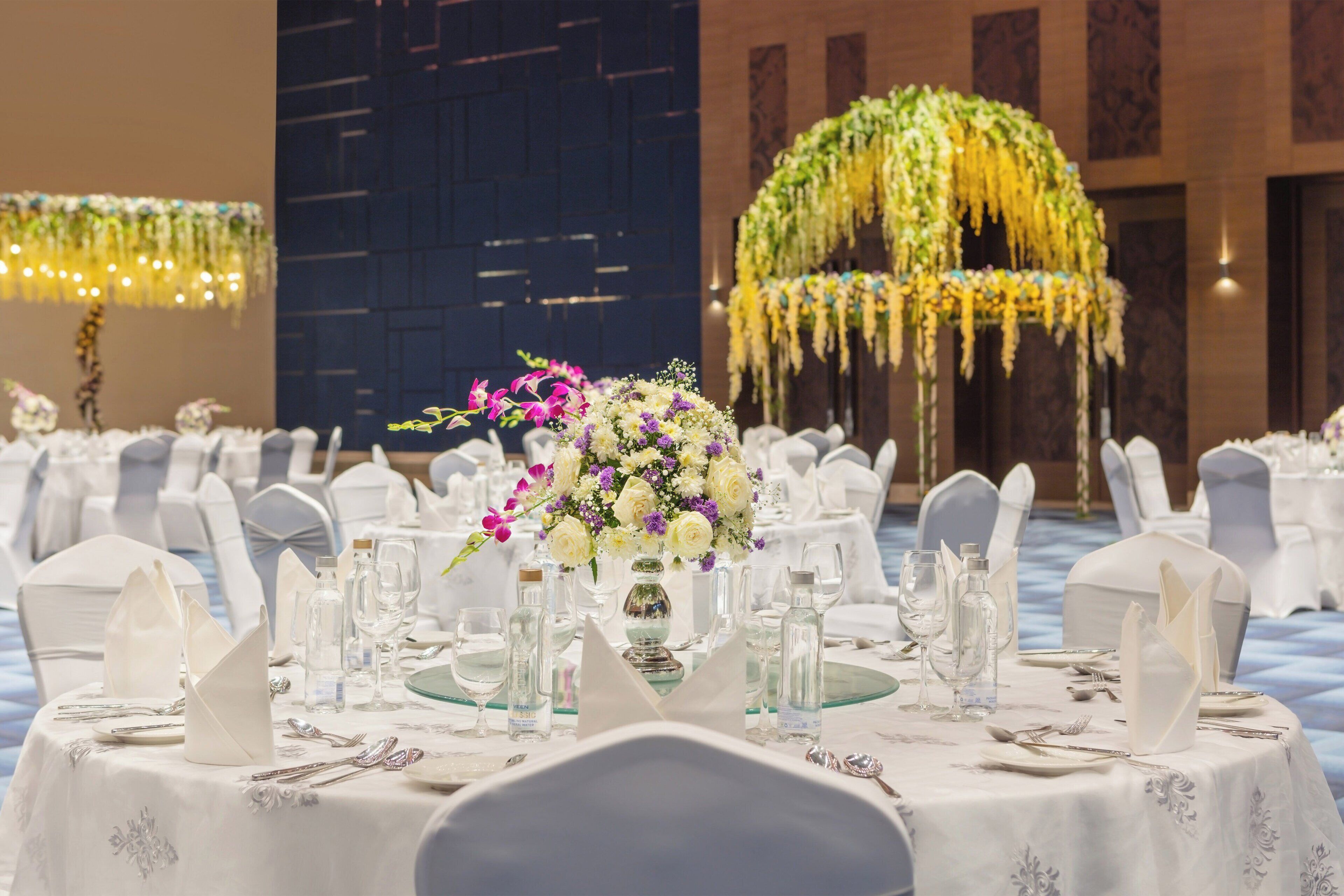Un salón de banquetes con mesas redondas dispuestas para un evento formal, con manteles blancos, cristalería y centros de mesa florales.