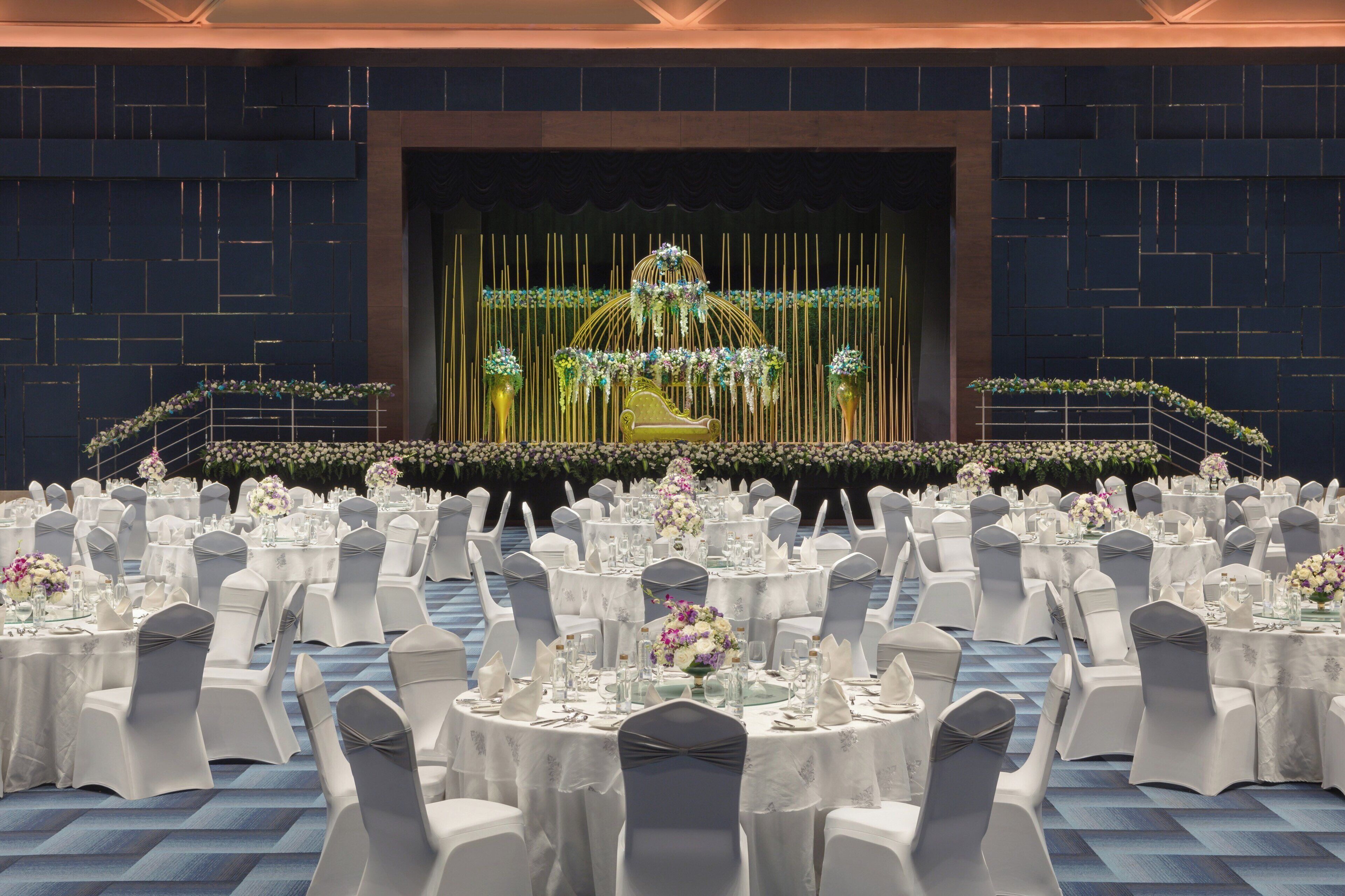 A banquet hall with round tables set for a formal event, featuring a stage with floral decorations and a golden chair.