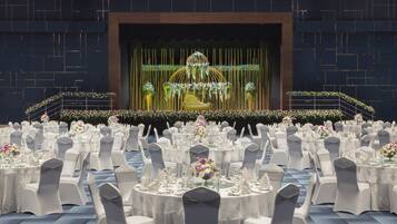 A banquet hall with round tables set for a formal event, featuring a stage with floral decorations and a golden chair.