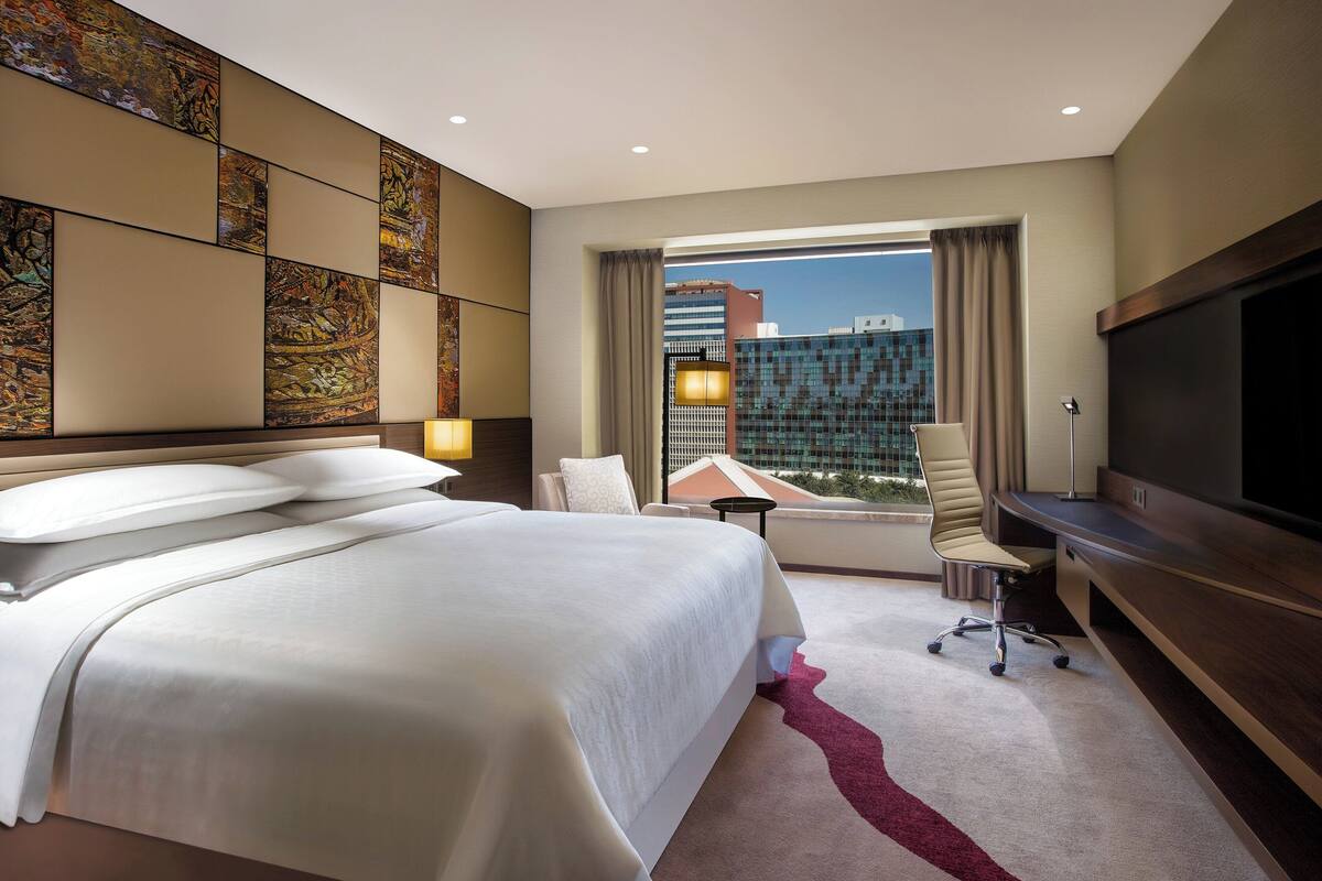 Habitación de hotel con una cama grande, un escritorio, una silla y un televisor. Se ve una ventana con vistas a edificios.
