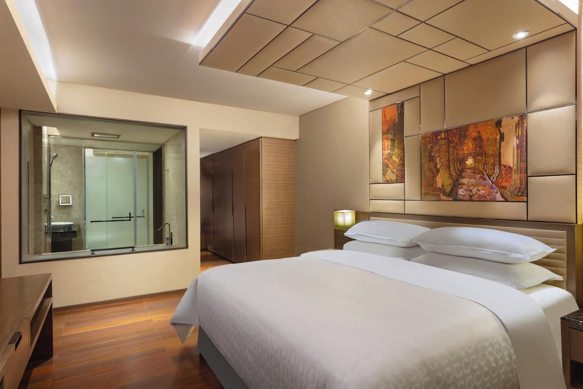 Una habitación de hotel moderna con una cama grande, ducha con cerramiento de vidrio, piso de madera y un cuadro abstracto de gran tamaño en la pared.
