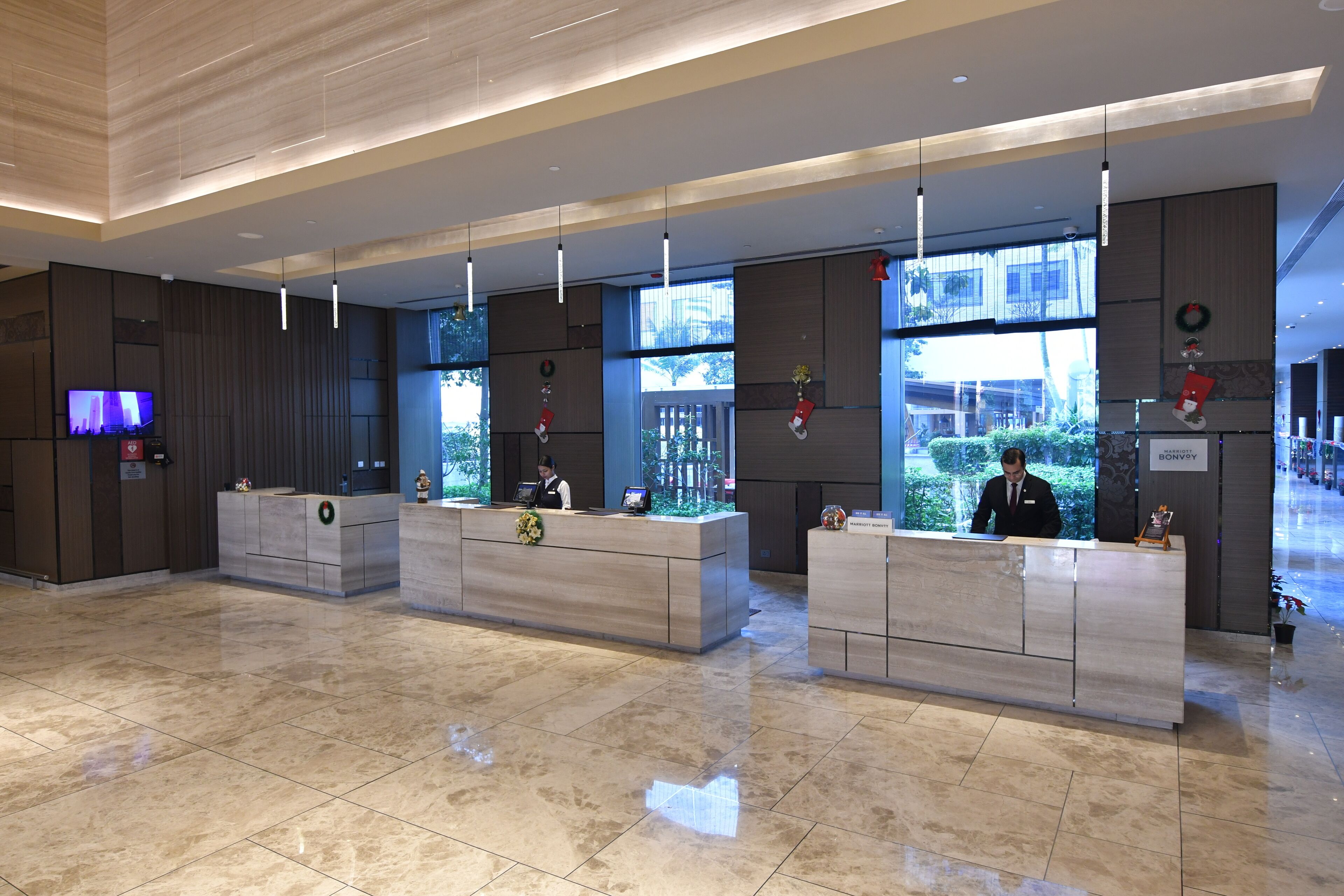 A modern hotel reception area with three reception desks, a flat-screen TV, and large windows offering views of the outdoors.