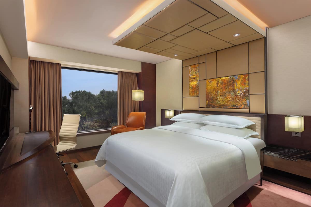 Una habitación de hotel moderna con una cama grande, un escritorio y una silla. Hay una ventana con cortinas y vistas a árboles.
