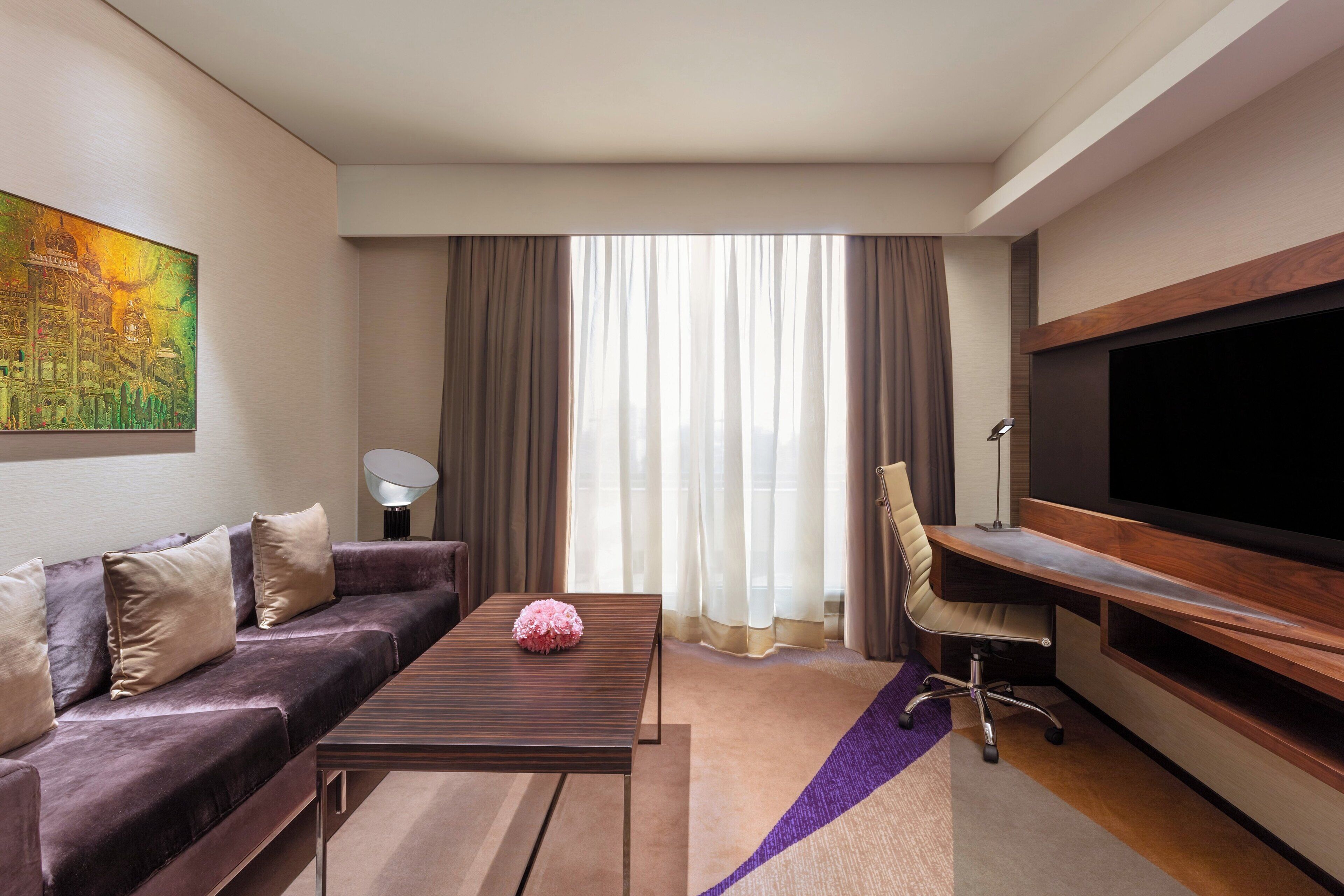A modern hotel room with a sofa, a coffee table, a desk, a chair, a flat-screen TV, and a large window with curtains.