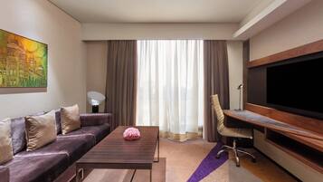A modern hotel room with a sofa, a coffee table, a desk, a chair, a flat-screen TV, and a large window with curtains.