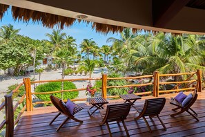 A wooden deck with lounge chairs and a table, overlooking a beach with palm trees and a clear sky.
