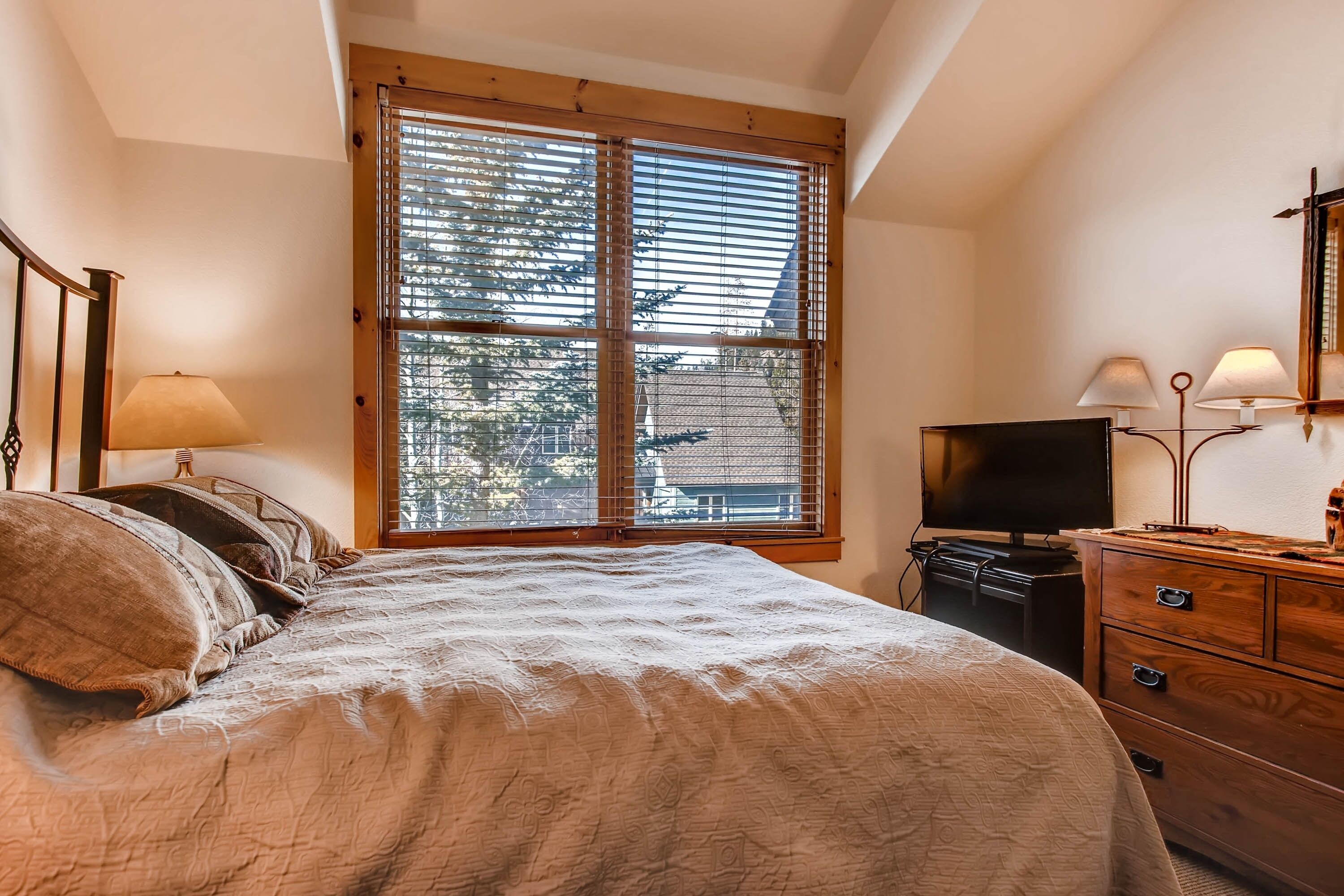 A bedroom with a large window, a bed, a dresser, and a television.