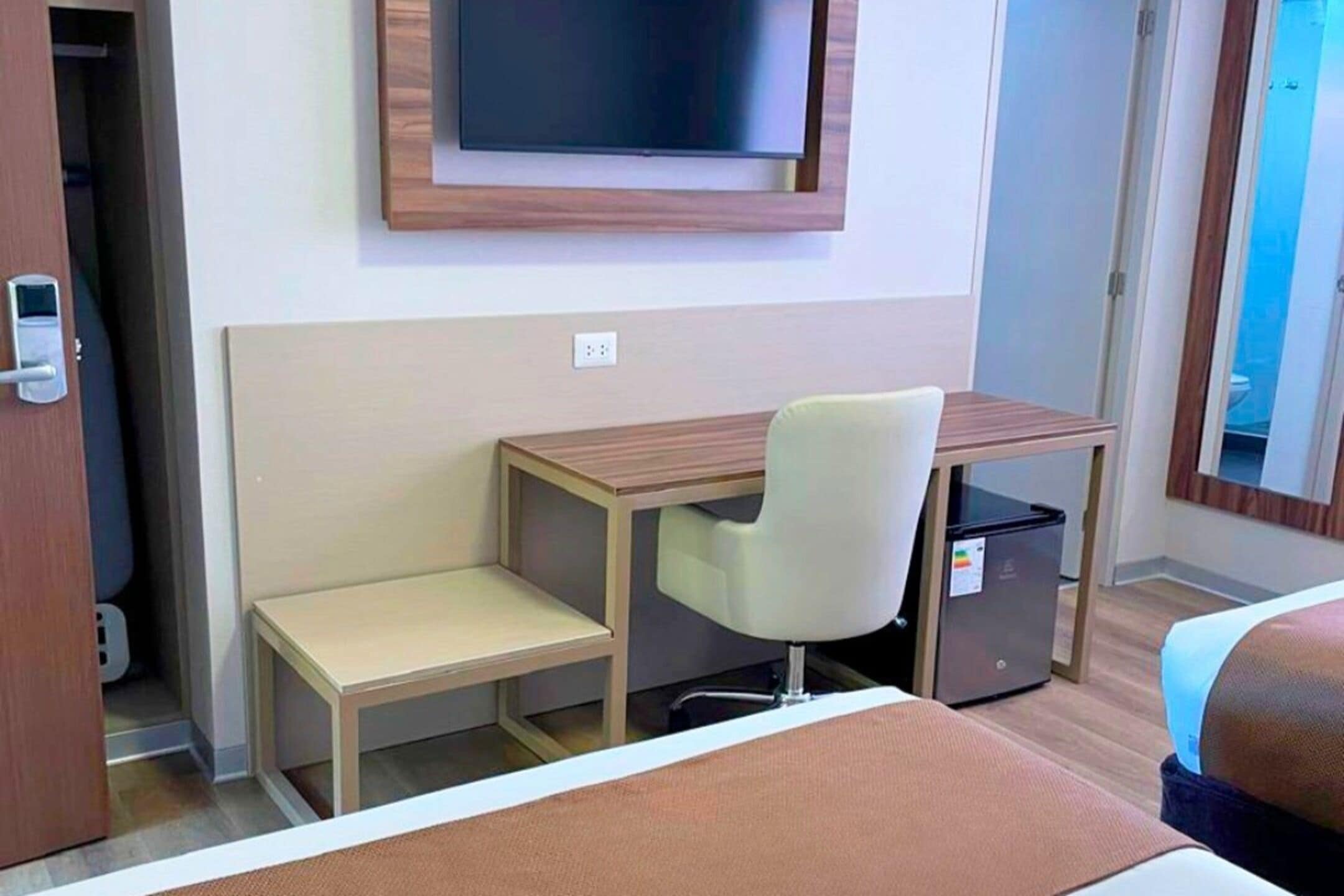 A hotel room with a bed, a desk with a chair, a television mounted on the wall, and a small refrigerator.