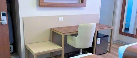 A hotel room with a bed, a desk with a chair, a television mounted on the wall, and a small refrigerator.