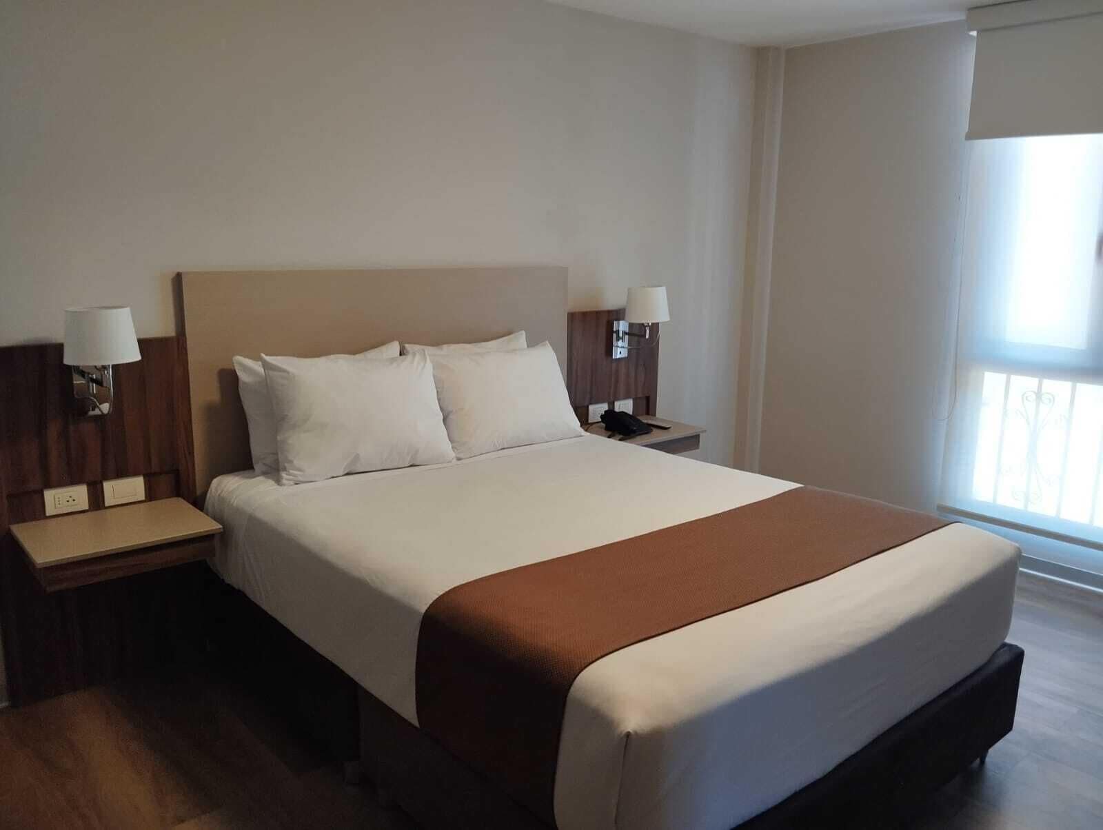 Una habitación de hotel con una cama grande, dos mesitas de noche y una ventana con persianas.